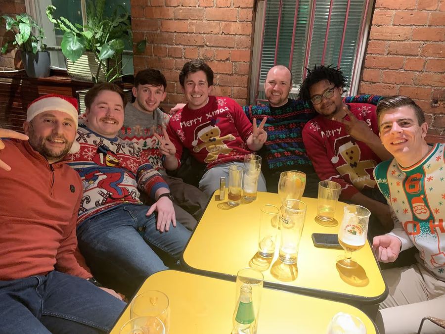 Kubicle teammates in Christmas jumpers around a yellow pub table, drinks raised.